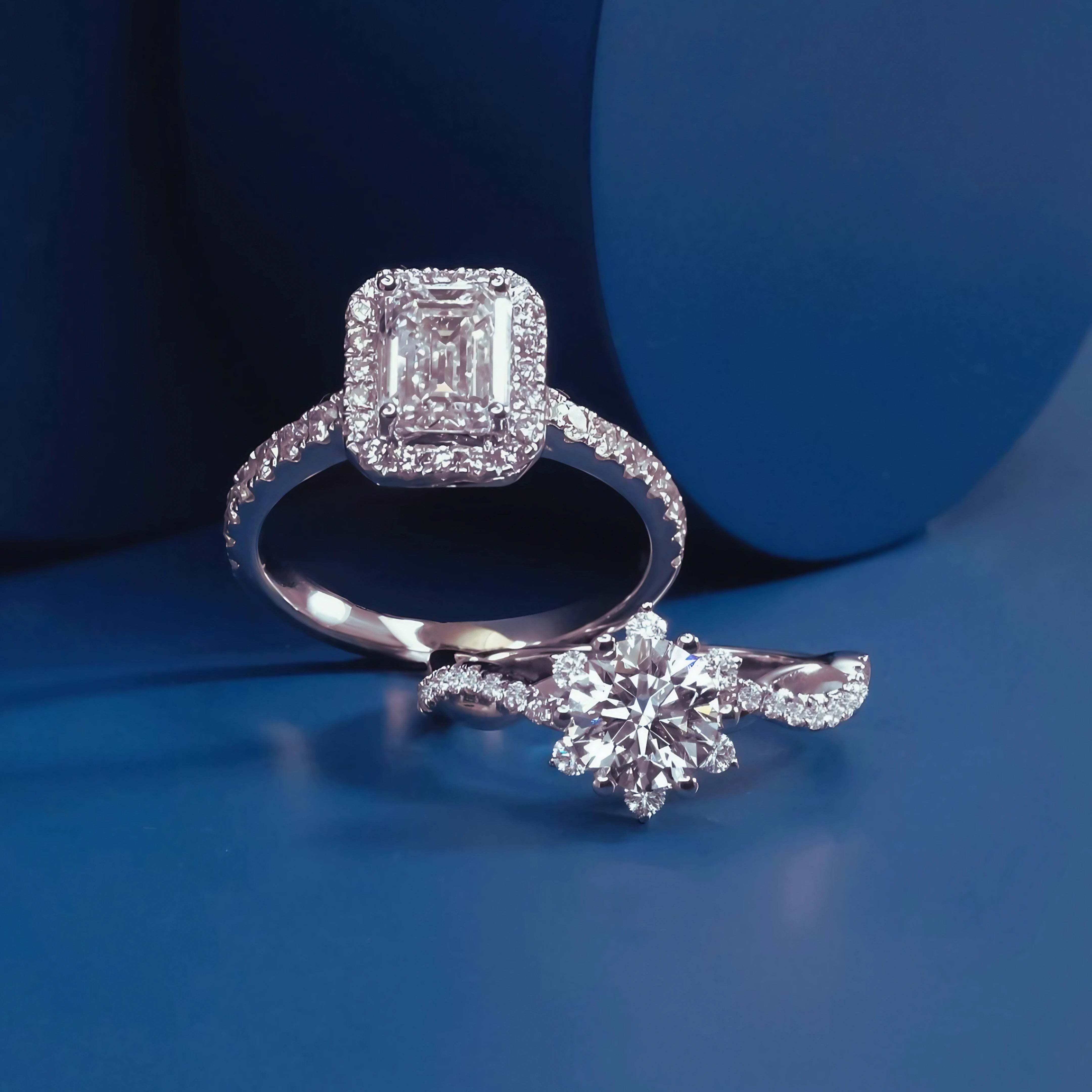 Two engagement rings on a matte blue backdrop, showcasing emerald and round cuts with pavé bands.