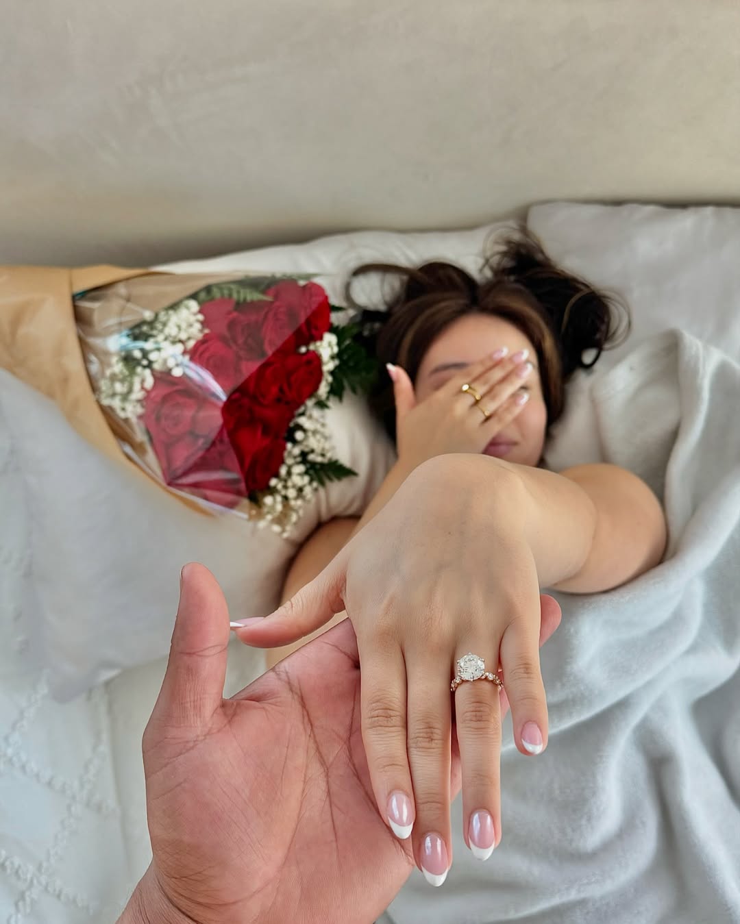 Romantic proposal scene in a bedroom with the woman lying on a bed holding flowers and showing her ring.