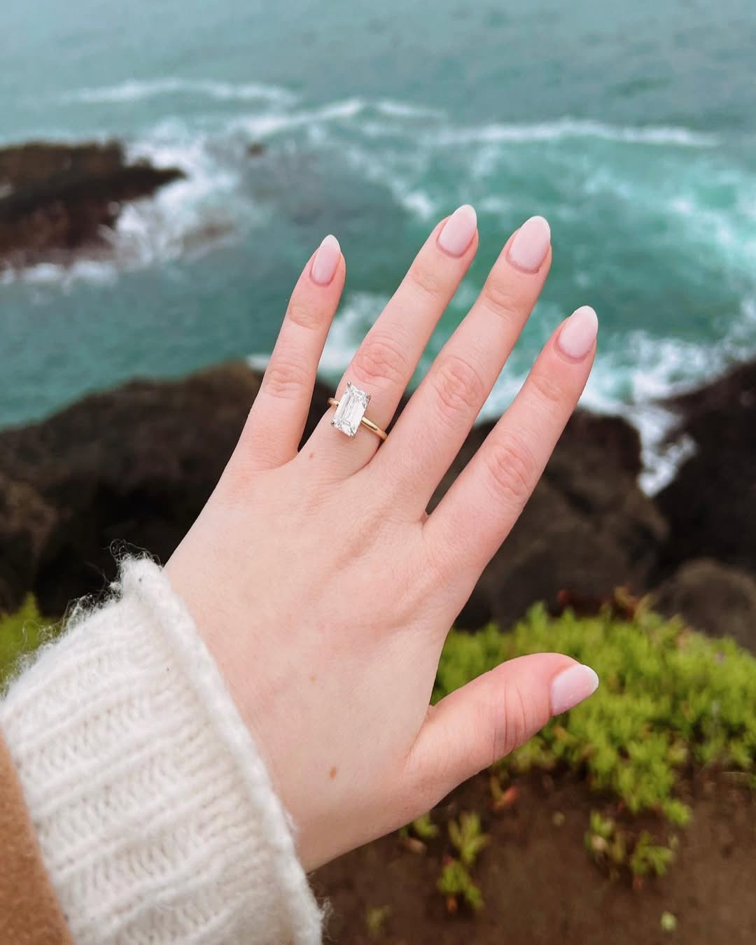 A hand raised toward the ocean with a solitaire engagement ring, capturing a breezy proposal moment.